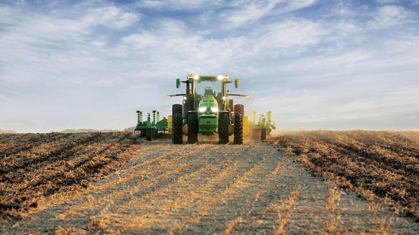 Itsekulkeva John Deere -traktori vetää maanmuokkauslaitteita aavalla pellolla.