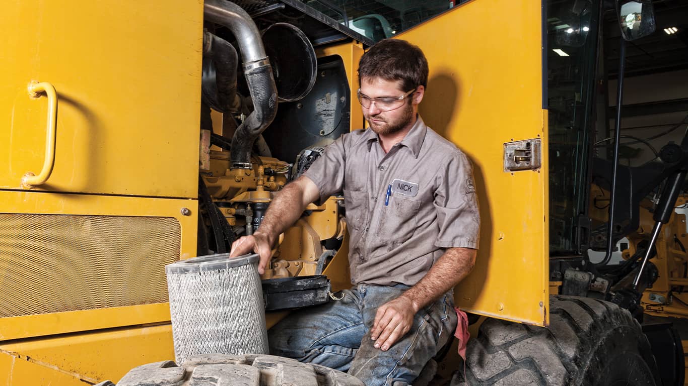 Suojalaseja käyttävä mies, joka työskentelee John Deere -moottorin parissa raskaaseen kalustoon kuuluvan koneen sisällä