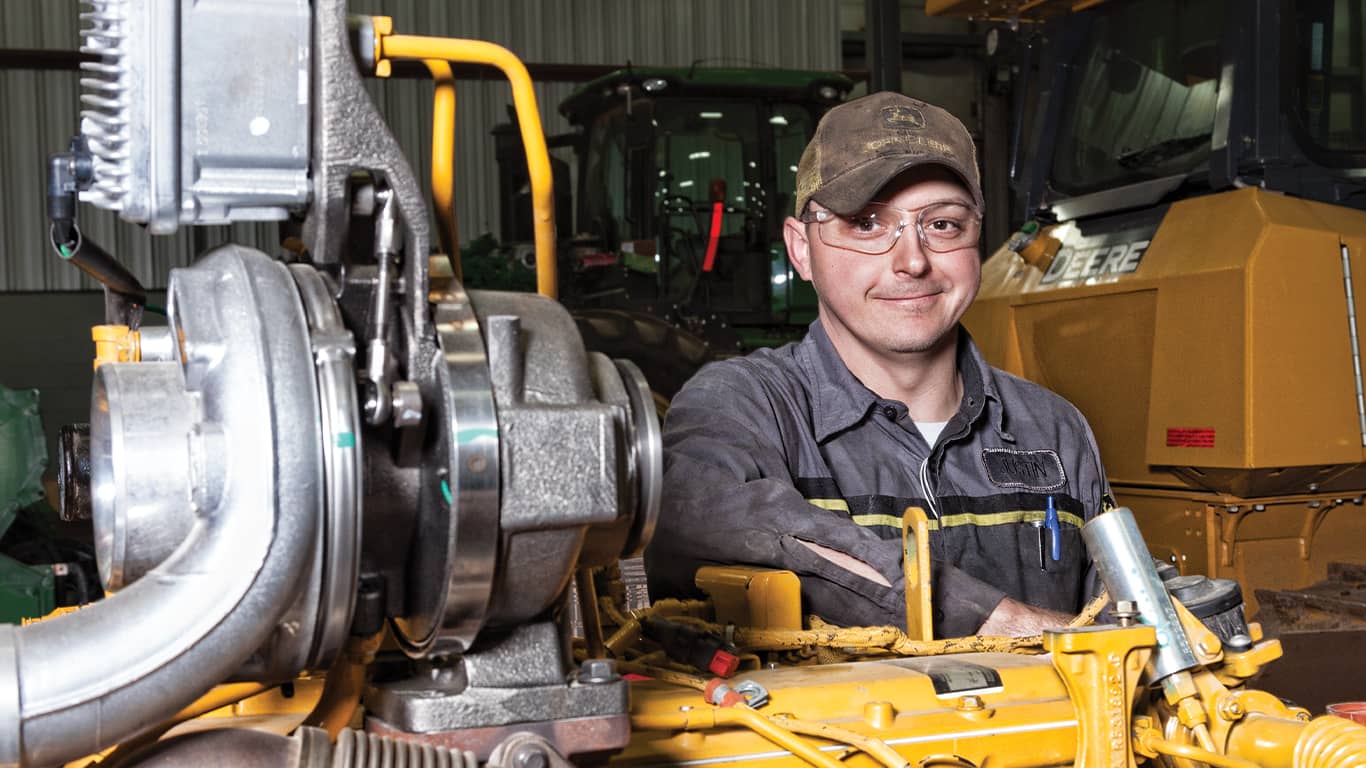 Suojalaseja käyttävä mies hymyilee John Deere -moottorin vieressä