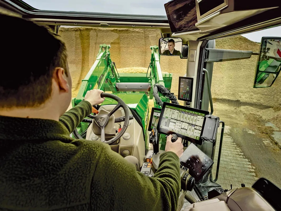 John Deere -traktorin ohjaamon sisänäkymä, jossa näkyvät kuljettajan hallintalaitteet ja etukuormaimen toiminta.