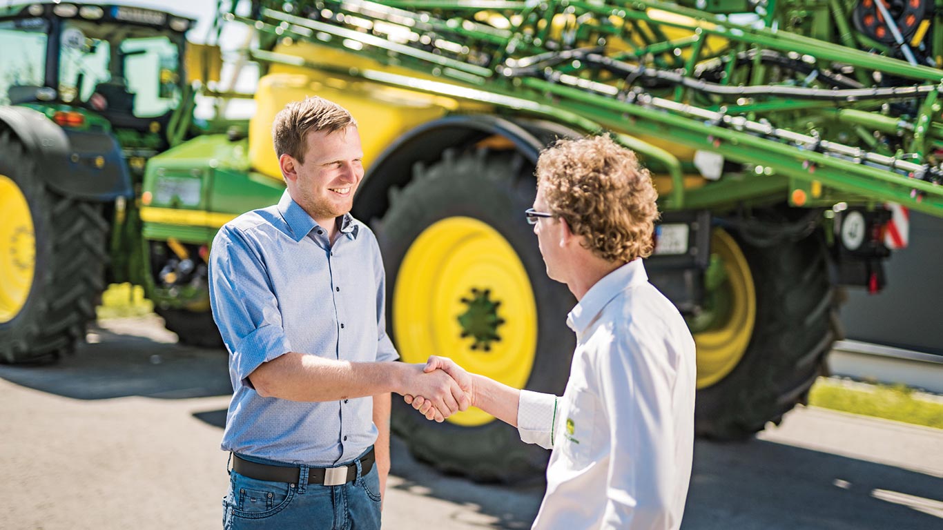 Jälleenmyyjä ja asiakas kättelevät John Deere -ruiskutuslaitteen edessä