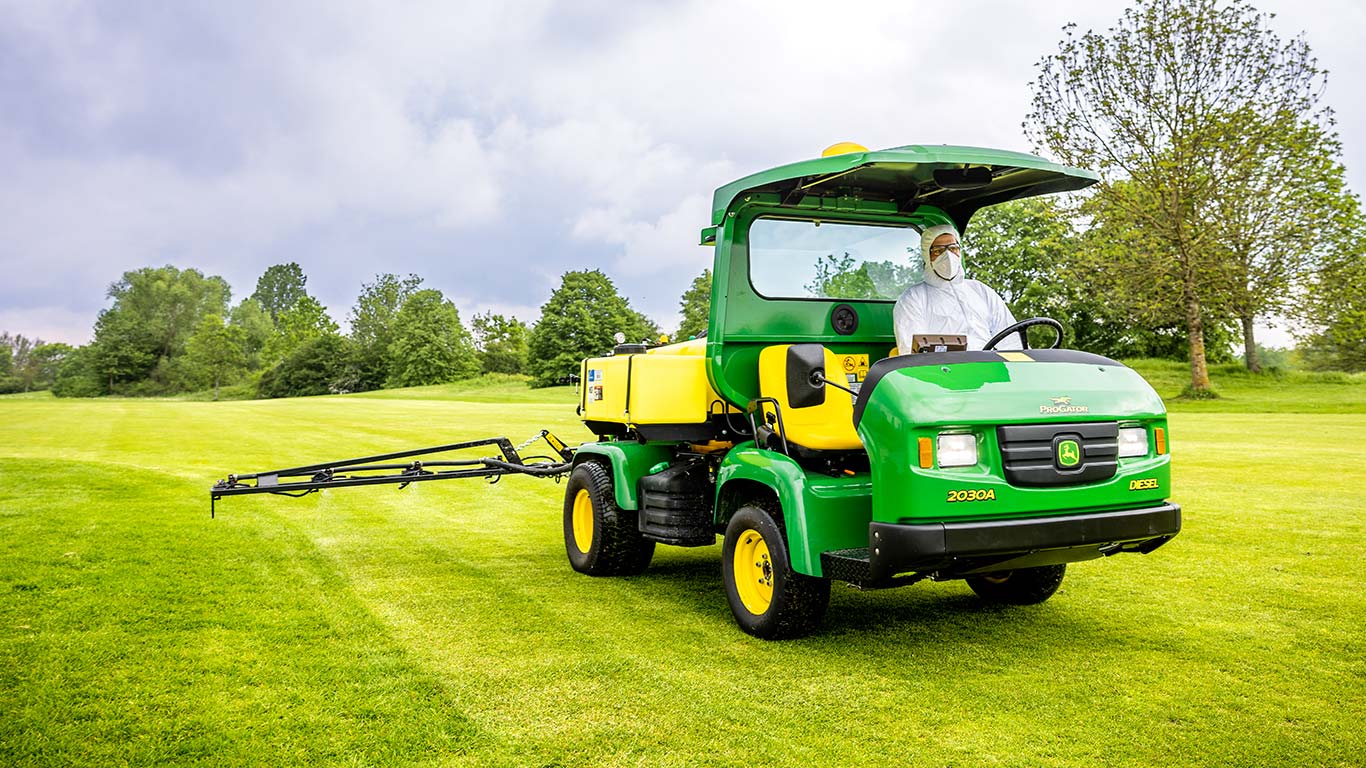 Suojavaatteisiin pukeutunut golfkentänhoitaja työskentelee golfkentän nurmella John Deere -ruiskutuslaitteella.