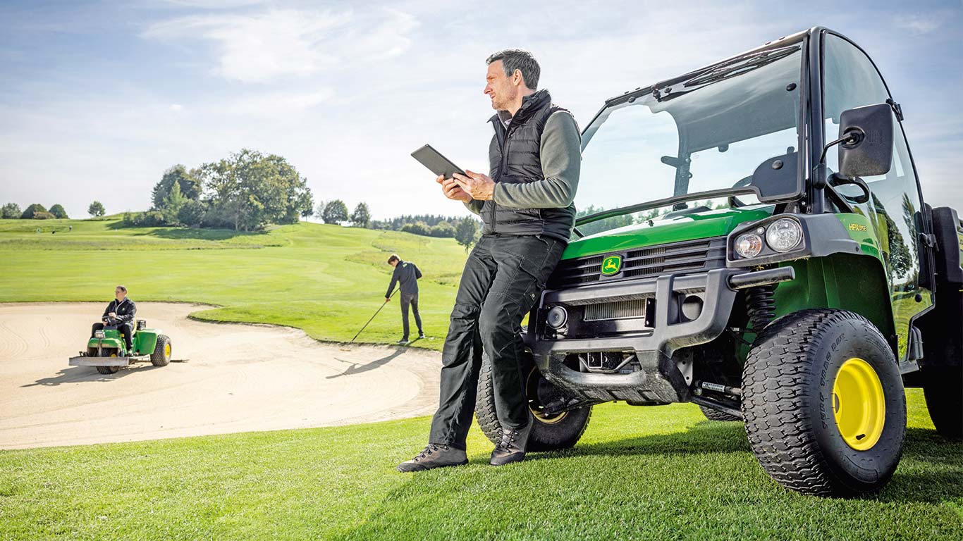 Vastaava golfkentänhoitaja tarkastaa tabletiltaan tietoja ja taustalla kaksi golfkentänhoitajaa työskentelee golfkentällä.