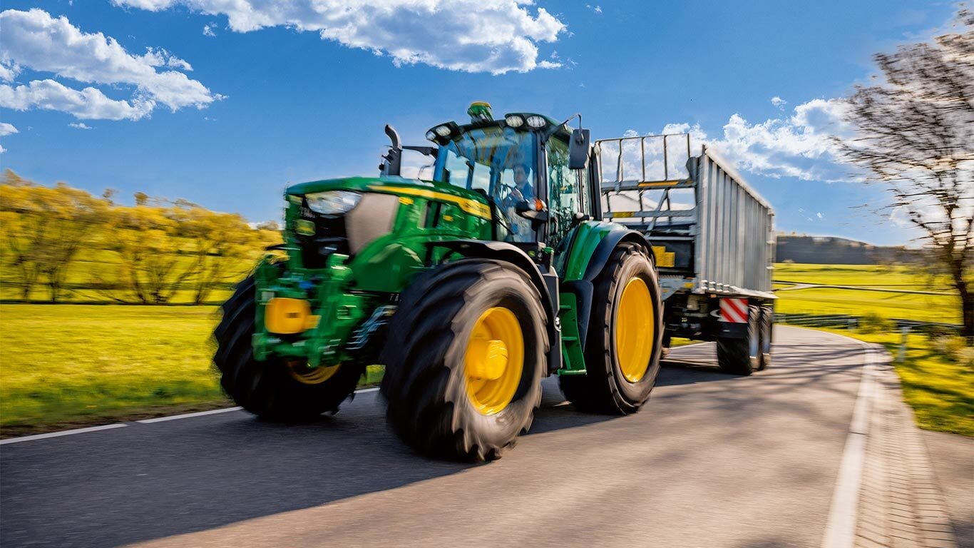 John Deere -traktori hinaa perävaunua maalaistiellä vihreällä maaseudulla sinisen taivaan alla puolipilvisellä säällä.