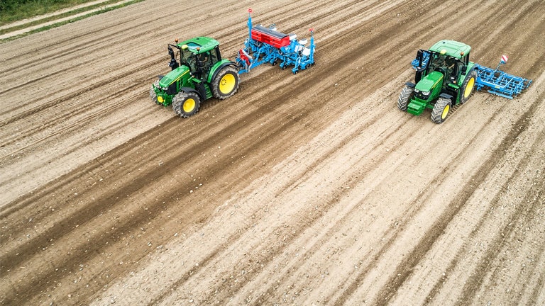 Kaksi John Deere 6M -traktoria paljaalla pellolla. Toinen vet&auml;&auml; Monsomen-kylv&ouml;konetta. Toinen vet&auml;&auml; Lemken-muokkausyhdistelm&auml;&auml;