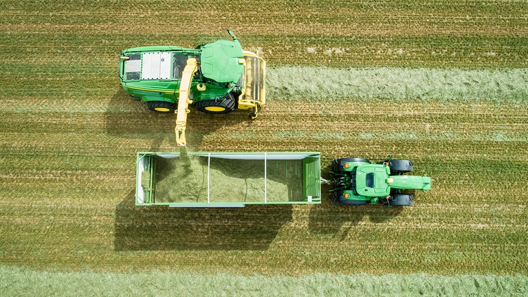 Ilmakuva John Deere 8000-sarjan itsekulkevasta ajosilppurista, jonka ruohonnoukin kuormaa s&auml;il&ouml;rehua John Deere -traktorin vet&auml;m&auml;&auml;n vaunuun