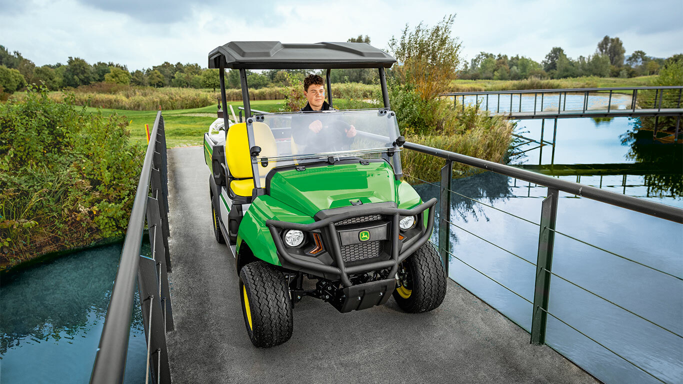 John Deere Gator GS -lava-ajoneuvo ajamassa kapeaa siltaa tyynen vesistön yli rehevän vehreyden ja puuston keskellä. John Deere Gator GS -lava-ajoneuvo ajamassa kapeaa siltaa tyynen vesistön yli rehevän vehreyden ja puuston keskellä.