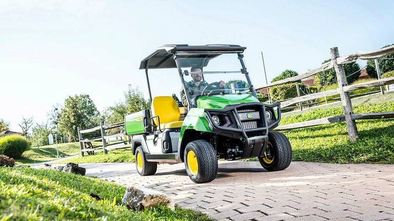 Vihreä John Deere Gator GS -lava-ajoneuvo ajamassa päällystetyllä tiellä maisemoidulla alueella, jossa on nurmikkoa ja puuaitoja.