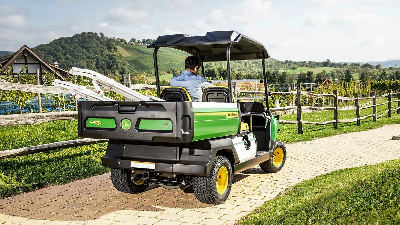 Takanäkymä John Deere Gator GS -lava-ajoneuvosta ajamassa kivetyllä tiellä taustalla luonnonkauniita kukkuloita ja viinitarhoja.