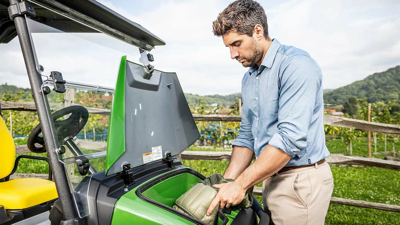 Henkilö asettamassa pehmustettua pussia vihreän John Deere Gator GS:n etusäilytyslokeroon ajoneuvon ollessa pysäköitynä ulkona puuaidan ja viinitarhan lähellä.