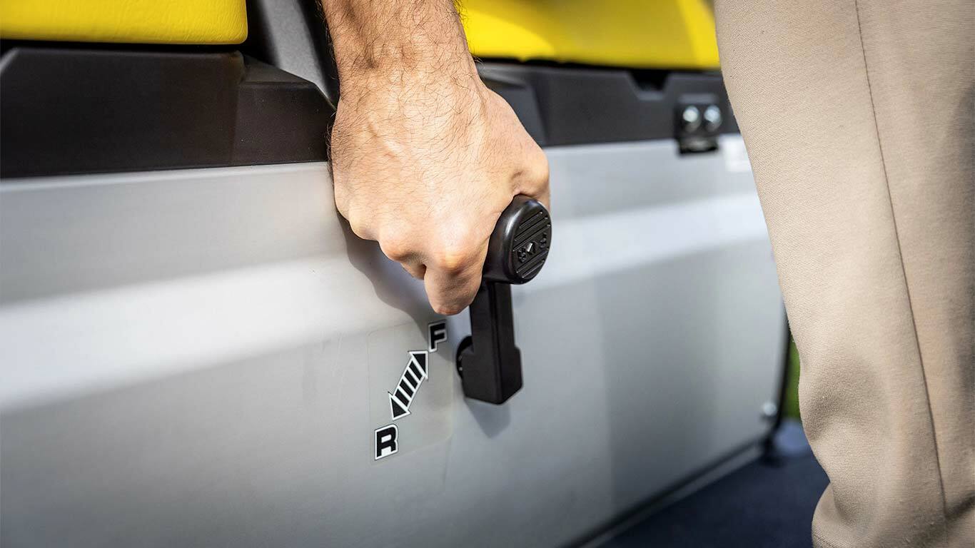 Lähikuva kädestä käyttämässä John Deere Gator GS:n vaihdevipua, jossa näkyvät eteenpäinajo- ja peruutusasennot, keltaisten istuimien vieressä.