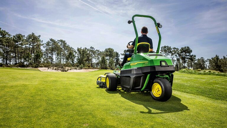 Henkilö ajaa vihreää sähkökäyttöistä John Deere 2775 E-Cut™ -viheriöleikkuria hyvin hoidetun golfkentän poikki, taustalla on puita ja osittain pilvistä taivasta.
