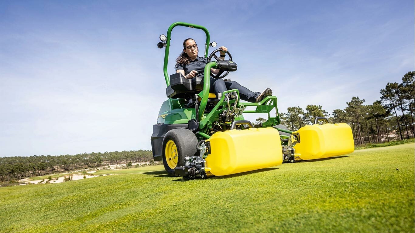 Henkilö ajaa golfviheriöllä viherkeltaisella sähkökäyttöisellä John Deere 2775 E-Cut™ -leikkuria, jossa on keltaisen kotelon peittämät eturullat, kun taustalla näkyy puita ja kirkas sininen taivas.
