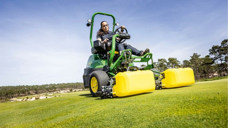 Henkilö ajaa golfviheriöllä viherkeltaisella sähkökäyttöisellä John Deere 2775 E-Cut™ -leikkuria, jossa on keltaisen kotelon peittämät eturullat, kun taustalla näkyy puita ja kirkas sininen taivas.