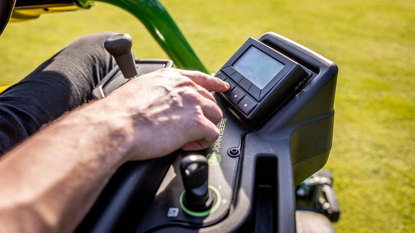 Lähikuva sähkökäyttöisen John Deere 2775 E-Cut™ -leikkurin TechControl-paneelia käyttävän henkilön kädestä, joka valitsee digitaalinäytön painikkeita joystick-ohjaimella, kun näkyvissä on myös muita hallintalaitteita.