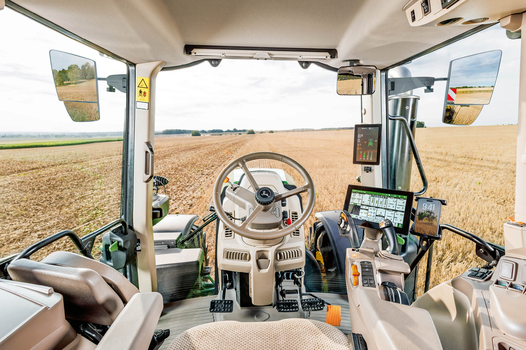 John Deere -traktorin ohjaamo sisäpuolelta, näkyvissä on ohjauspyörä, digitaaliset näyttölaitteet ja peltonäkymä tuulilasin läpi maatilan töiden aikana.