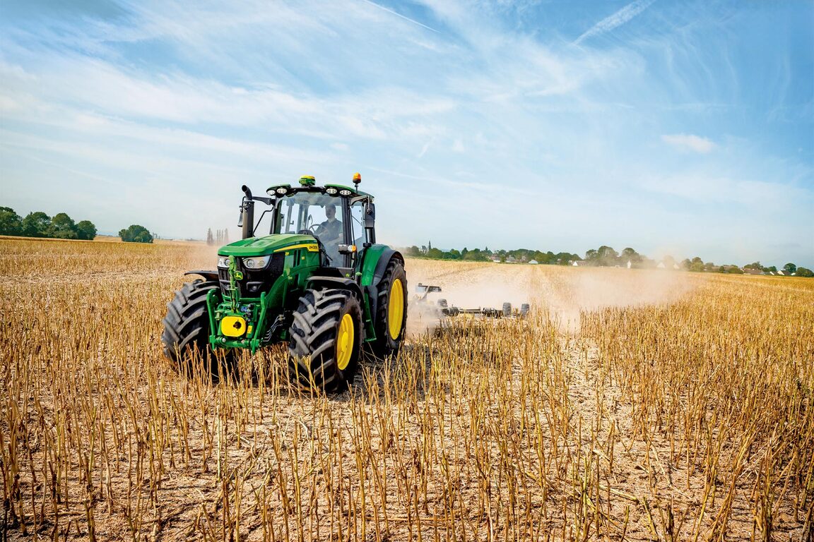 John Deere -traktori pellolla sadonkorjuun aikaan. Pölyä nousee ilmaan, kun se vetää maatalouskoneita kirkkaansinisen taivaan alla.