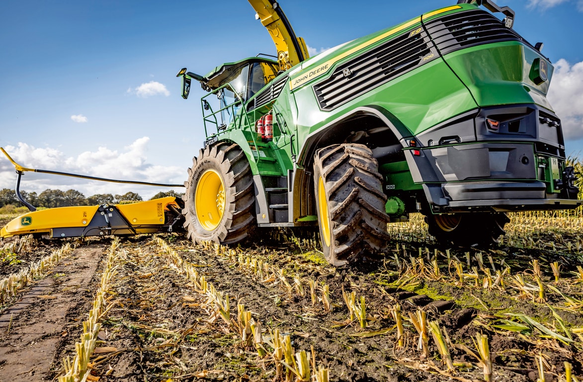 John Deere -ajosilppuri työskentelee korjatussa maissipellossa, esittelee koneen suuria renkaita ja leikkuumekanismia kirkkaan sinistä taivasta vasten.