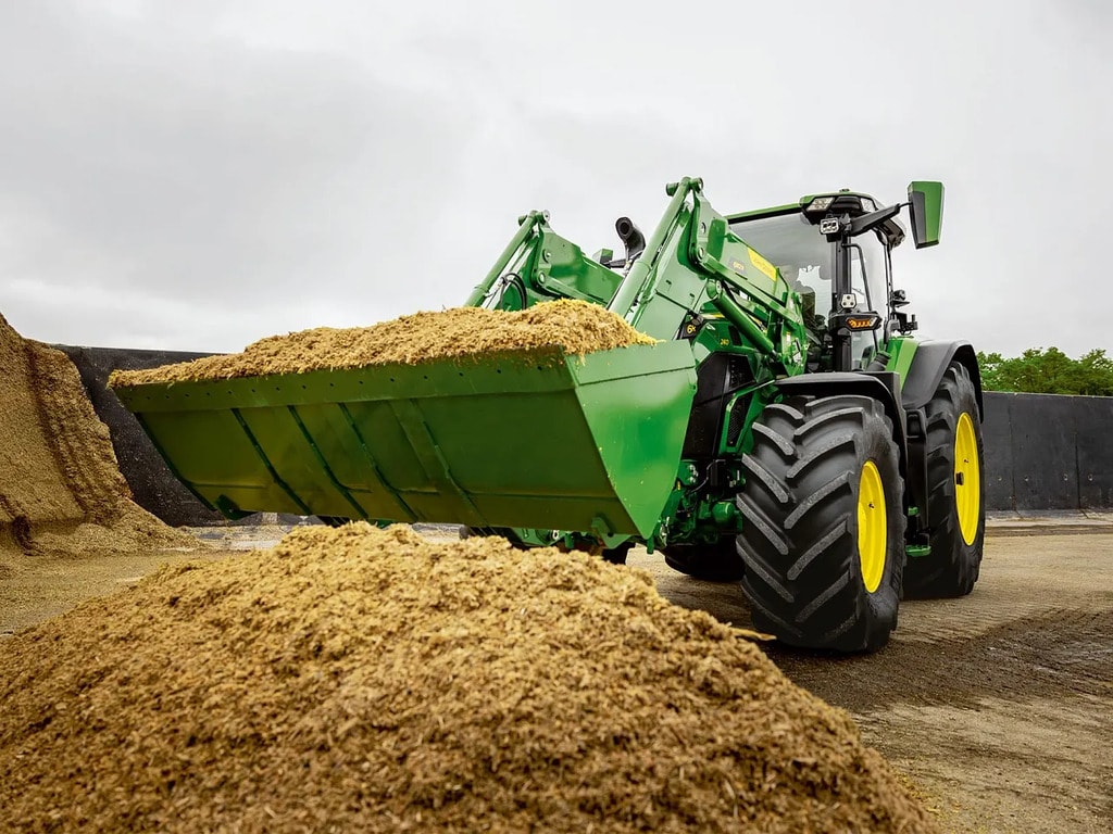 John Deere -traktori kuljettamassa silputtua säilörehua etukuormaimella maatilan varastoalueella.
